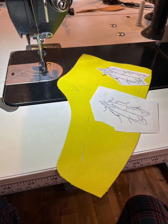 Two hand-sketched moths with closed wings on cutout paper,
tacked to a yellow leather high-top side panel,
on the sewing machine.