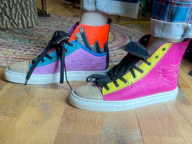 Low angle me wearing the shoes,
right foot inside has lavender low-top with blue lining
that transitions to silver/black stripes and orange upper.
Left foot outside is magenta with yellow lace panel.
Both have a mix of tan stitched art,
black stitched horizontal lines,
and red stitched scribbles.