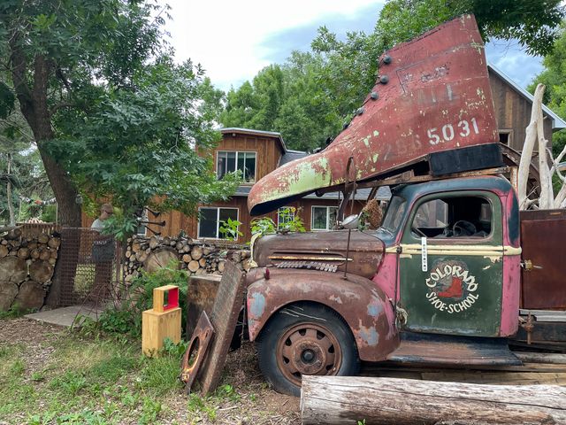 A rusting vintage pickup
with Colorado Shoe School painted on the door
and a massive metal boot mounted on the cab.
Immediately behind that,
a fence of stacked wood with ends showing,
and Erin standing behind a small metal gate under a tree.
A house and barn in the background.