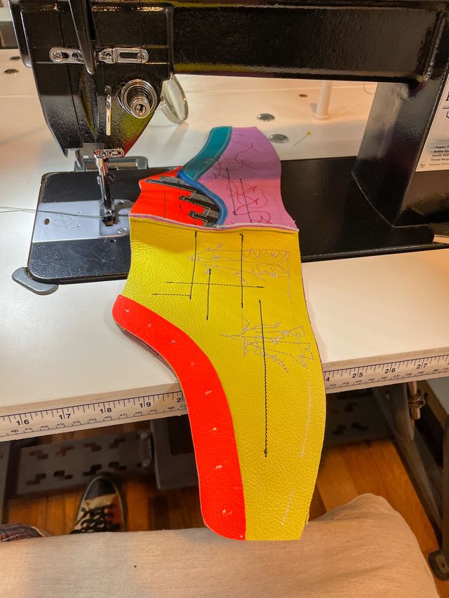 On a sewing machine,
the yellow high-top is sewn at the heal
to a lavender low-top and orange addition.
The yellow side has stitching of two moths,
and some straight black lines across,
with orange lacing panels.
The lavender side has a blue trim
and then silver-and-black stripes protruding
up and forward into the orange addition.