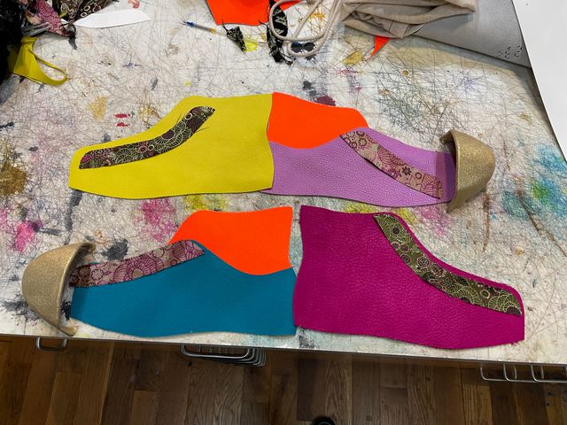Top view cut-out parts for both shoes
laid out on the work table.
Top shoe has yellow high-top outer,
and lavender low-top inner,
with orange high-top addition.
Bottom shoe has magenta high-top outer,
with cyan low-top inner
and same orange high-top addition.
Both have the same floral lace panel,
and gold leather toe-caps.
