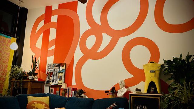A blue couch with yellow pillows, an old console tv converted to hold records, various artworks on the counter, plants, and a white wall painted with two orange pillars and large lighter orange swirls that match the fabric of a yellow & orange curtain.